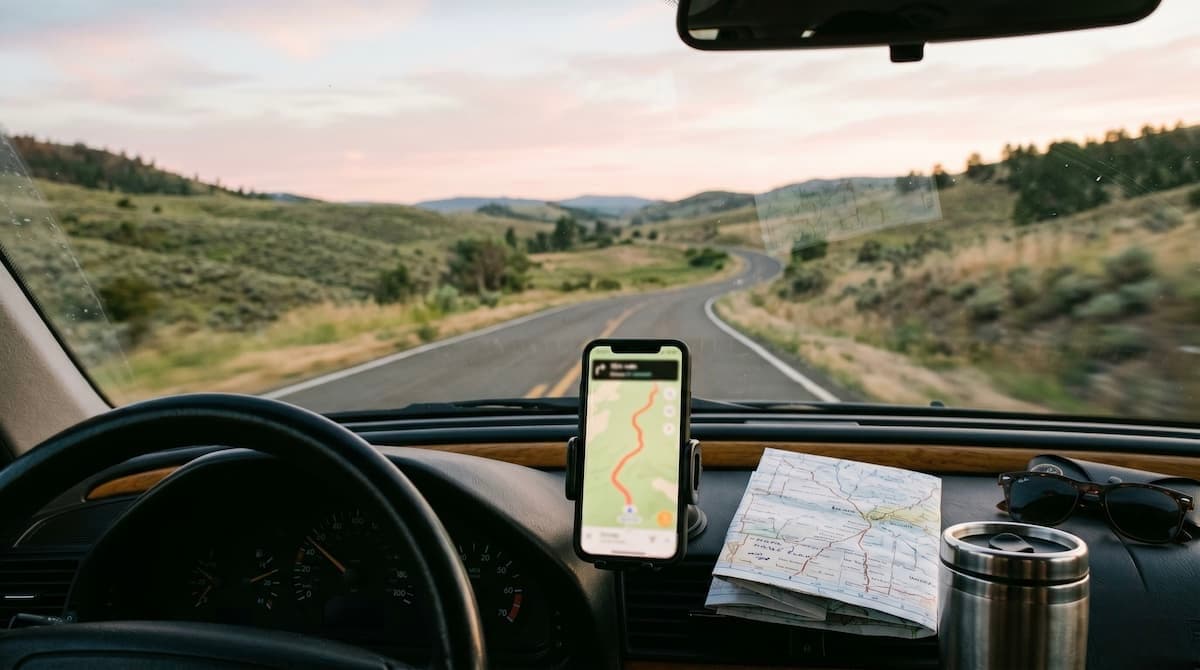 A dashboard-perspective view through a windscreen onto a winding country road at golden hour, with a phone mounted on the dash showing a navigation map