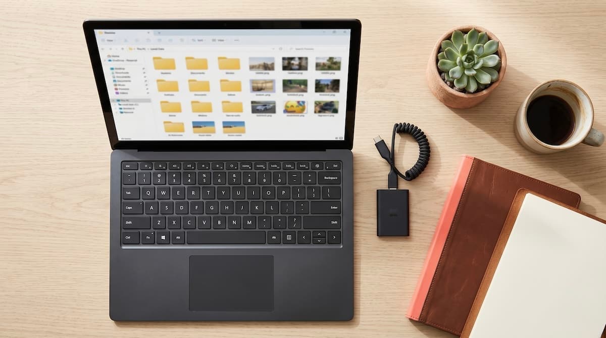 An overhead view of a matte laptop on a pale oak desk showing organised folders and photo thumbnails, with an external SSD, notebook, and coffee alongside